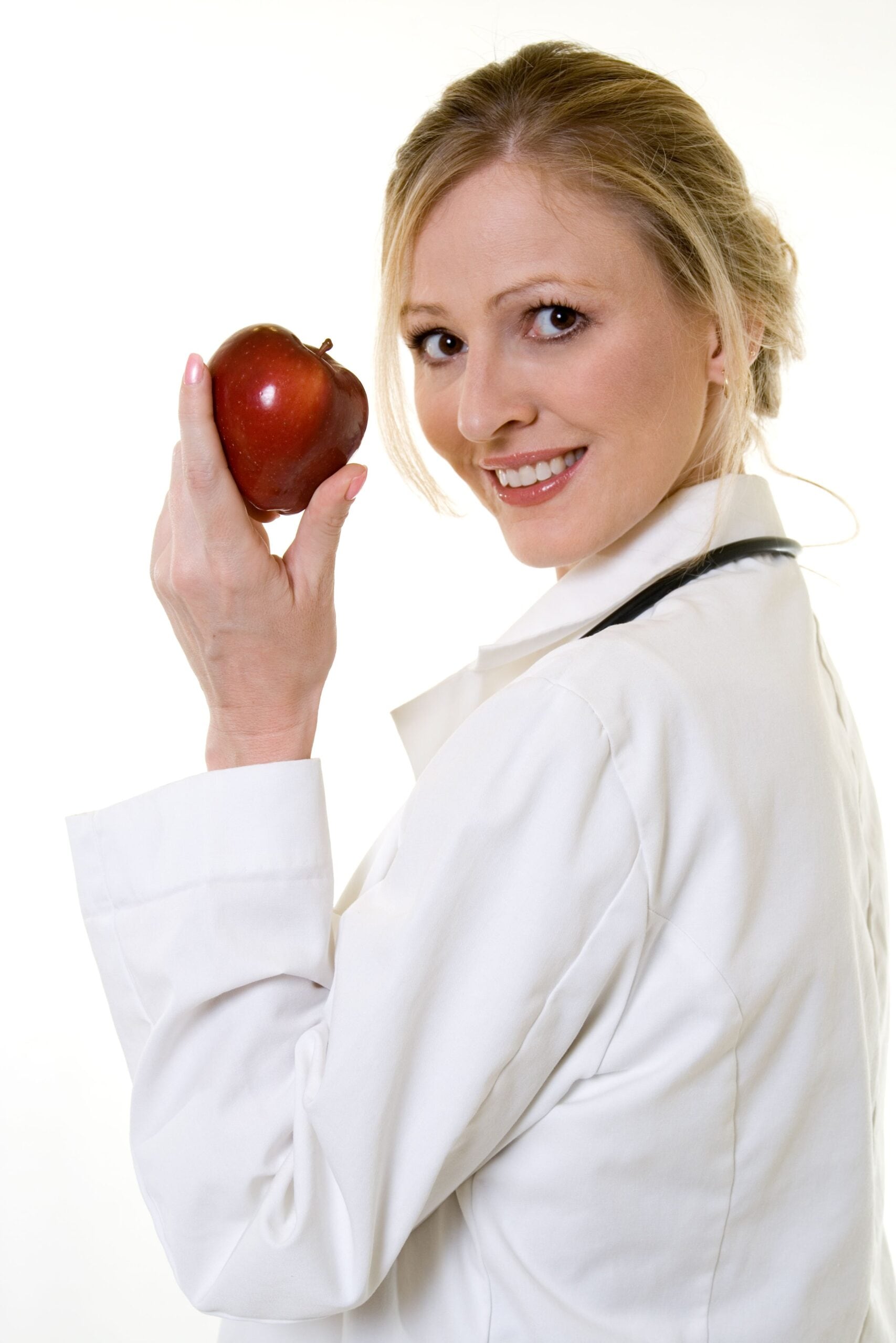 a woman with blond hair wearing a white doctors coat and holding a red apple looks over her shoulder at the camera and smiles