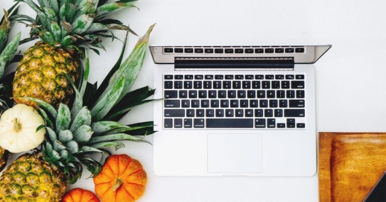 Laptop surrounded by fresh pineapples, apples, and small pumpkins on a white surface.
