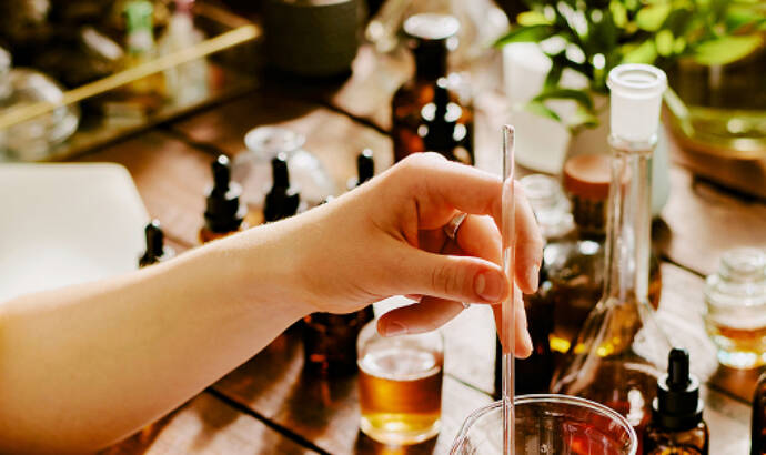 Hands measuring amber essential oil into a glass beaker on a scale, surrounded by dropper bottles on a wooden table