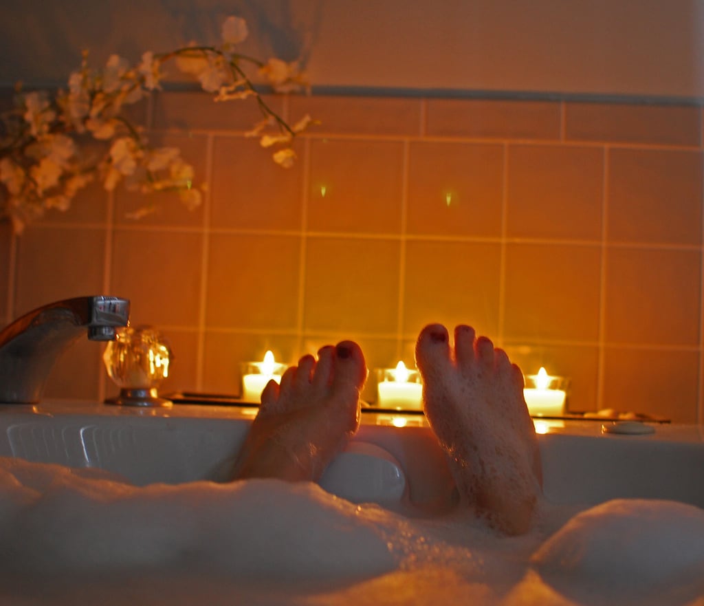 two feet with red painted toenails stick out of a bubble bath with candles behind them