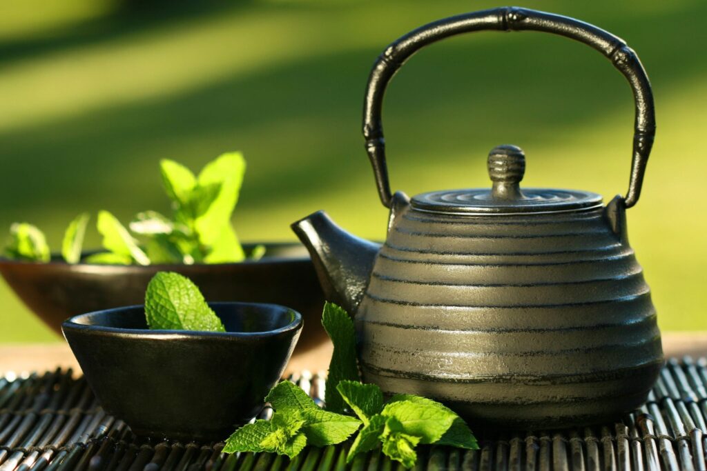 a cast iron teapot sits on a bamboo mat next to a small black teacup, tea and mint leaves are in the cup and in the background