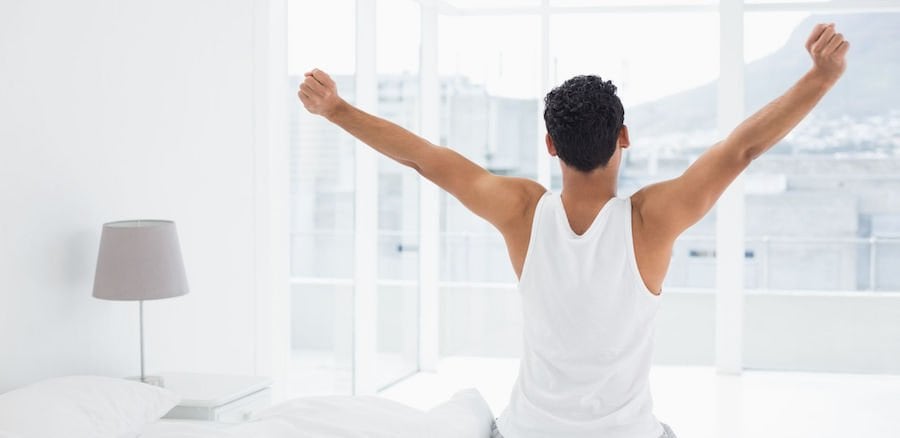 man in a white tank top stretches his arms to his sides as he sits on a white bed in a white room, looking out a floor to ceiling window