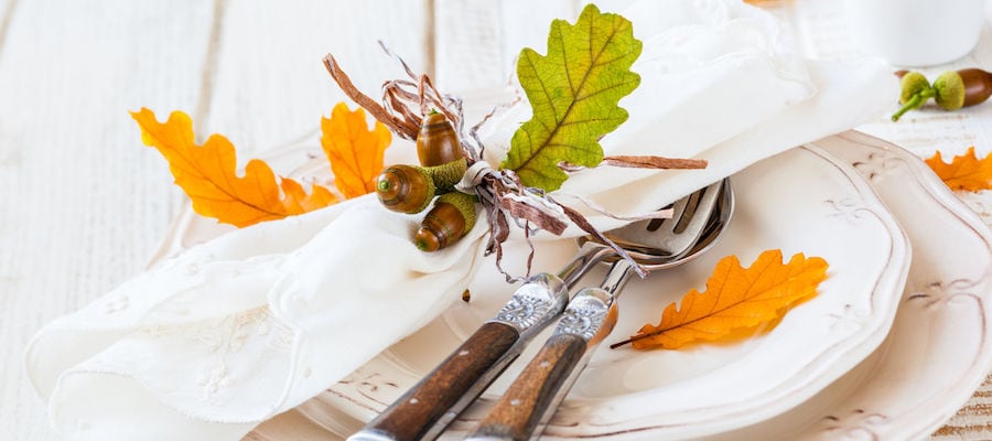 a fall tablscape including white plates, silver and wooden silverware and a white napkin wrapped in ribbon and decorated with leaves and acorns