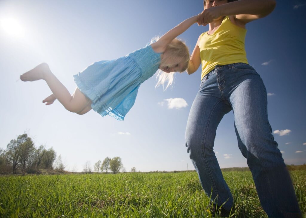 an adult woman swings a child in a blue dress around in an open field with green grass