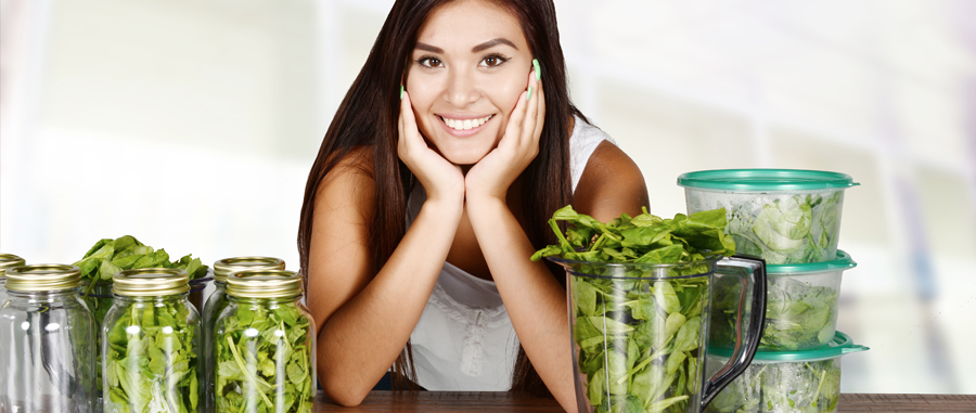 a woman with dark hair leans on a table with her elbows. the table holds jars and containers of leafy greens