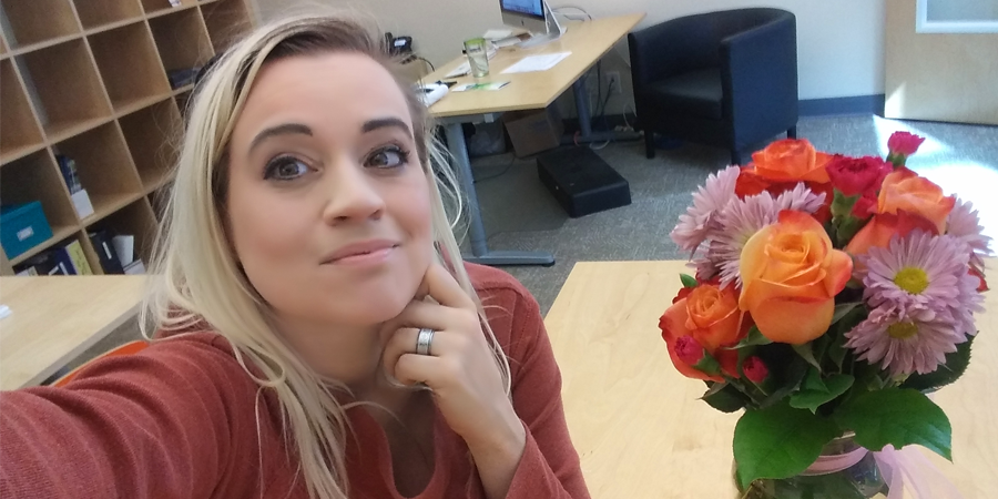 blonde woman takes her own photo next to a bouquet of flowers in an office setting