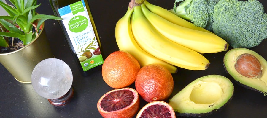 fruits and veggies are laid out on a dark table, including bananas, avocado and broccoli