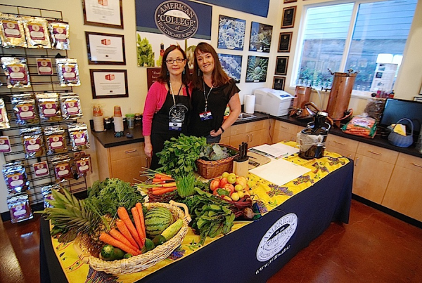 two women with long, dark hair stand together in a kitchen with a table covered in fruits and veggies