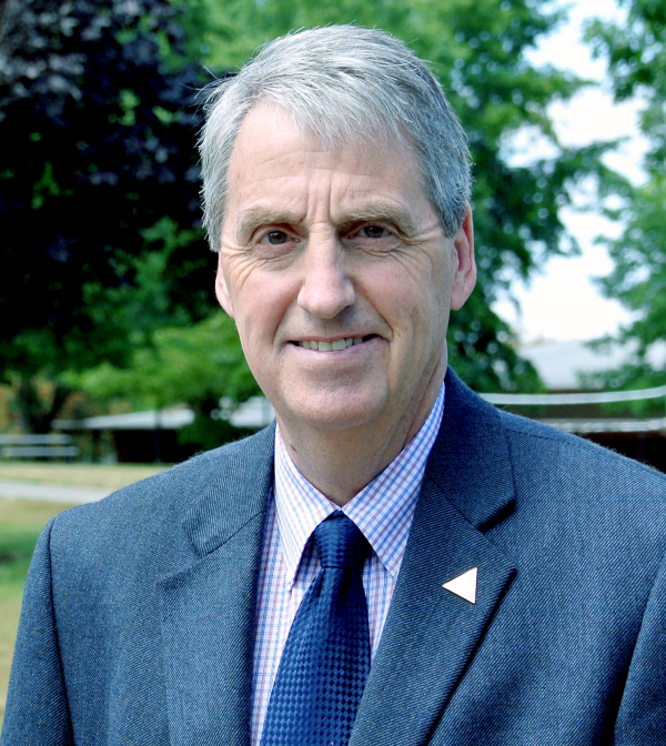 man with grey hair wearing a blue suit jacket over a checkered collared shirt and blue tie smiles at the camera