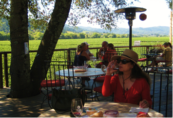 a woman in a red top and straw hat sips wine at a round table under a tree in an outdoor restaurant setting