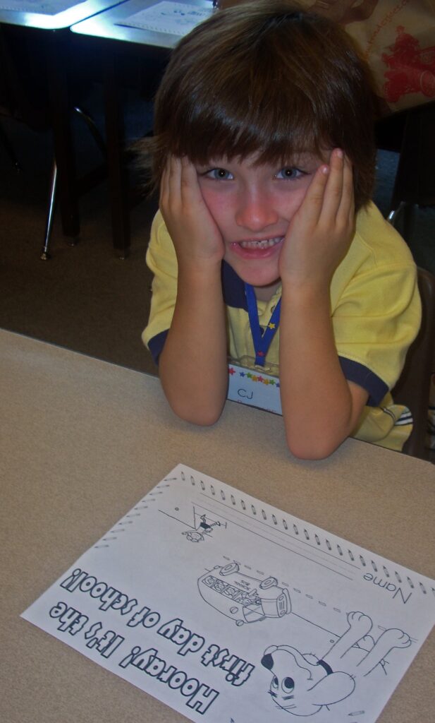 a child with dark brown hair holds their face in their hands and looks at the camera whilst their elbows are on the desk in front of them