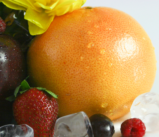 close up shot of the outside of a grapefruit with water droplets on it and a strawberry, blueberry and raspberry in front of it