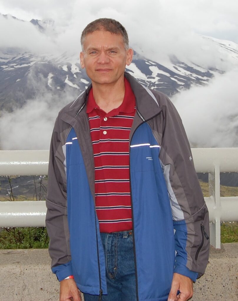 man with short hair, red striped shirt and blue and grey jacket stands outside in front of a smoky mountain skyline