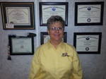 a woman in a yellow long-sleeve shirt smiles at the camera with six certificates hanging on the wall behind her