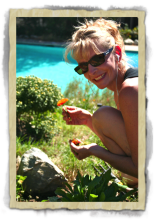 blonde woman in sunglasses leans down to pick an orange flower, looking to her side and smiling at the camera