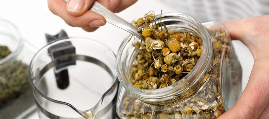 a person holds a glass jar of dried chamomile with one hand and scoops it out using a silver spoon with the other hand