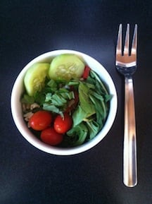 a white ramikan is filled with greens, tomatoes and cucumber slices, a silver fork sits next to it on the table