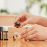 Hands pouring essential oil into a glass bowl, with small amber bottles of essential oils on a wooden table in the background.