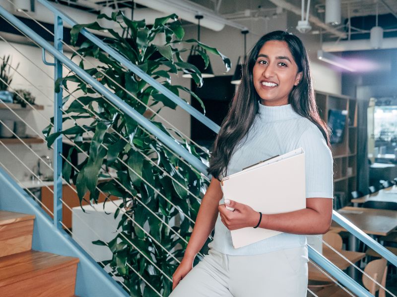 A smiling woman holding a binder stand on a staircase in an office setting