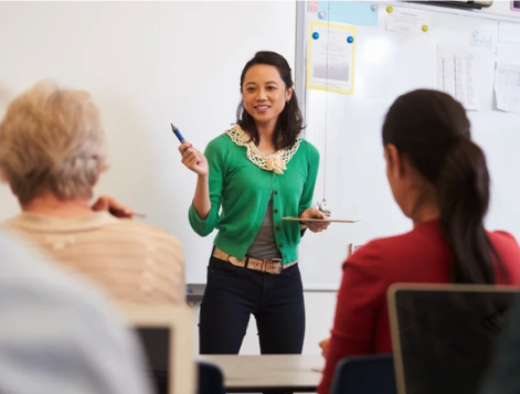 A woman in a green sweater stand at the head of a classroom teaching
