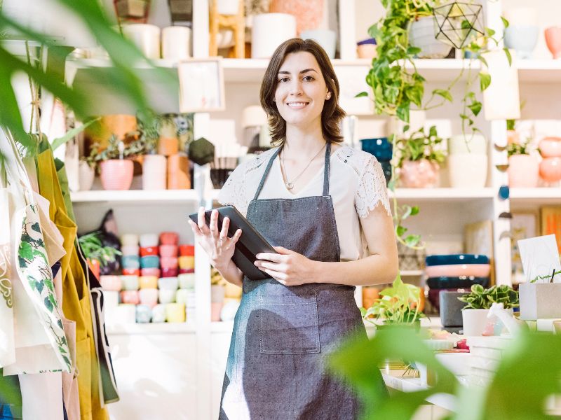 A woman wearing an apron and holding an iPad stands inside a small business