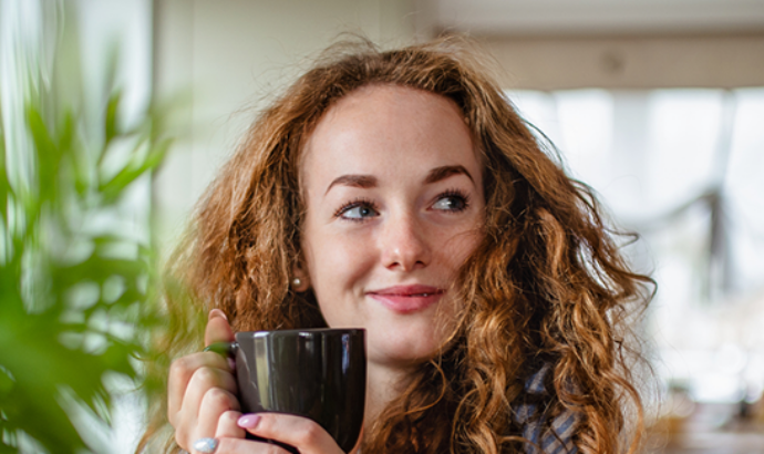 Woman with curly hair holding a mug while surrounded by potted plants indoors.