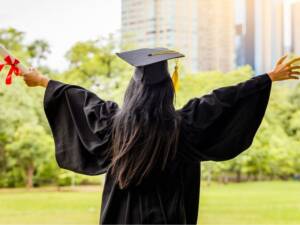 A graduate in regalia holding a diploma with both hands in the air