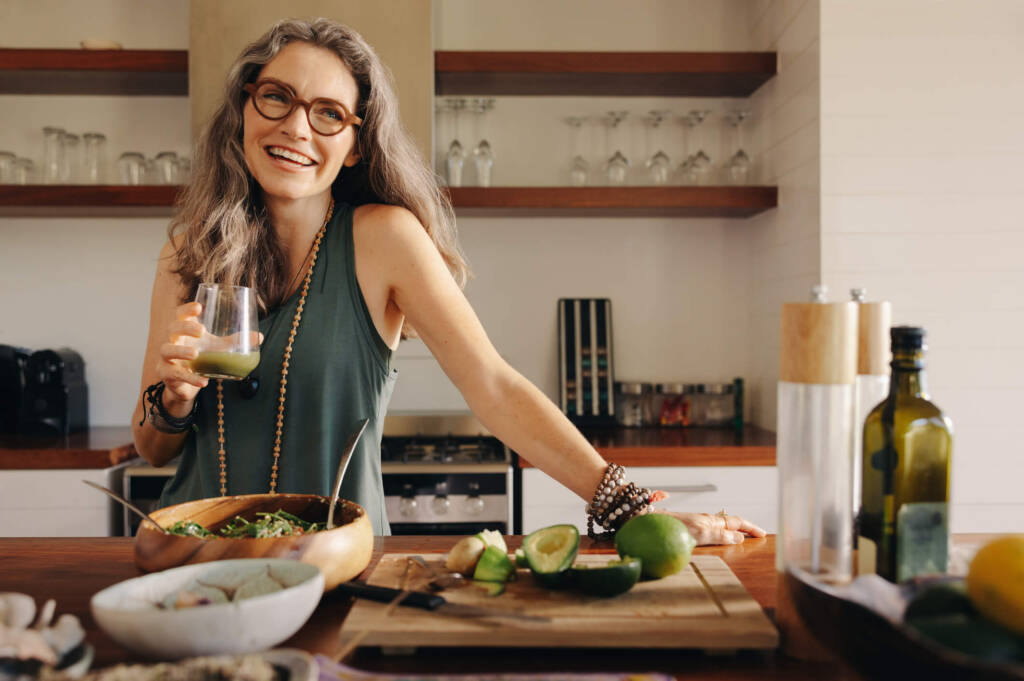 Smiling woman holding a green smoothie in a kitchen with avocados, limes, and a salad bowl on the counter
