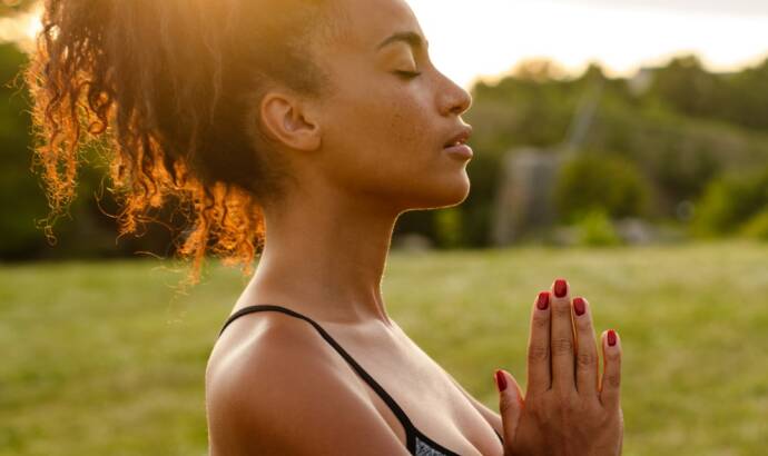 A woman stands outside on grass at sunset, eyes closed and hands pressed together in a prayer pose, wearing a sports bra and leggings.