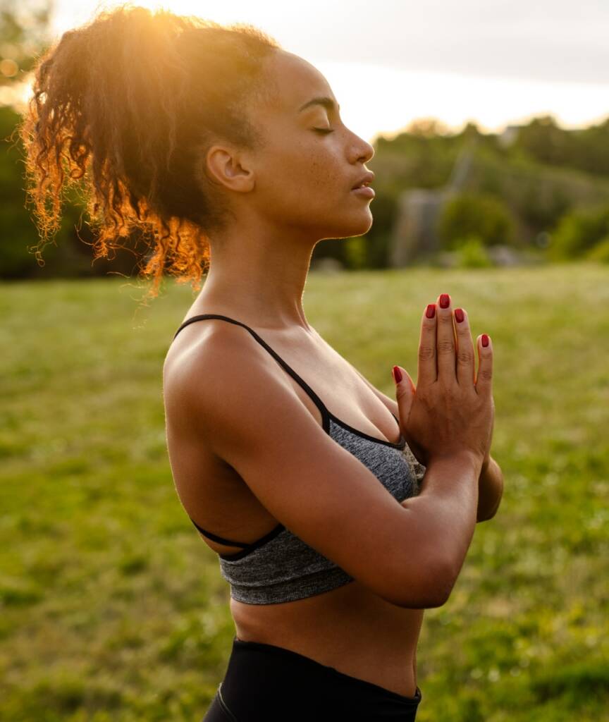 A woman stands outside on grass at sunset, eyes closed and hands pressed together in a prayer pose, wearing a sports bra and leggings.