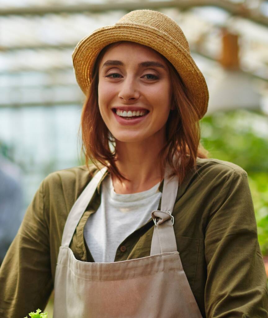 Smiling woman wearing an apron and straw hat in a garden setting.