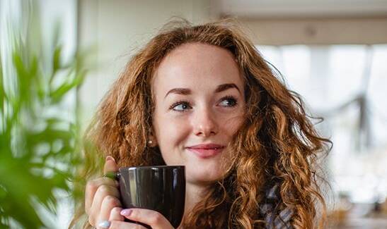 Woman with curly hair holding a mug while surrounded by potted plants indoors.