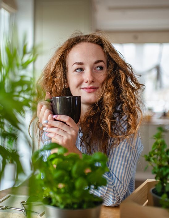 Woman with curly hair holding a mug while surrounded by potted plants indoors.
