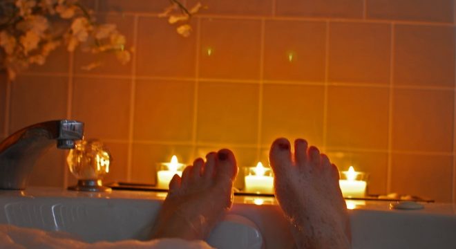 two feet with red painted toenails stick out of a bubble bath with candles behind them