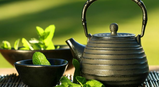 a cast iron teapot sits on a bamboo mat next to a small black teacup, tea and mint leaves are in the cup and in the background