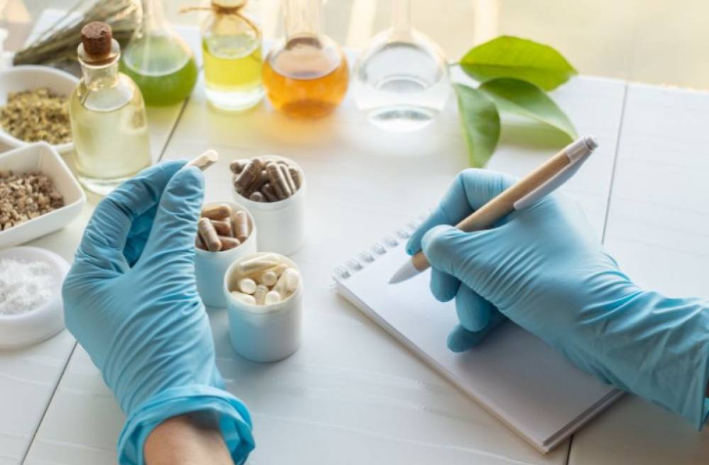 Person wearing blue gloves examines capsules and writes notes beside herbal ingredients and laboratory glassware.