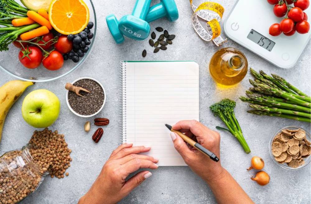 Person writing in a notebook surrounded by fruits, vegetables, whole grains, olive oil, weights, and a body weight scale, representing healthy lifestyle planning.