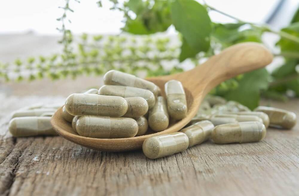clear pills with a brown/green powder sit on a wood table and in a wooden spoon on the table