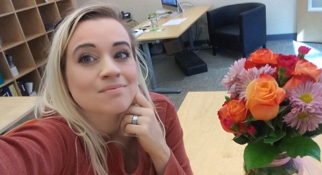 blonde woman takes her own photo next to a bouquet of flowers in an office setting