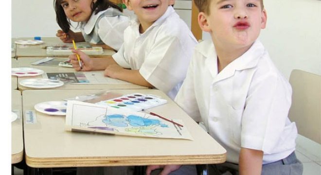 three grade school children sit at their desks, all wearing white and looking and smiling at the camera as they paint