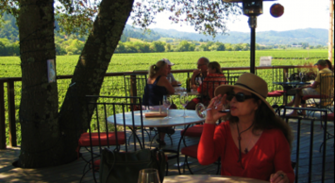 a woman in a red top and straw hat sips wine at a round table under a tree in an outdoor restaurant setting