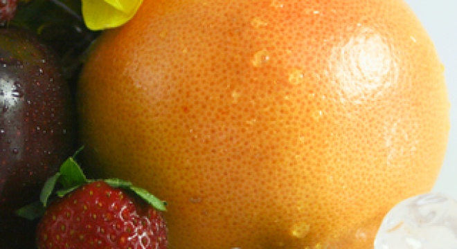 close up shot of the outside of a grapefruit with water droplets on it and a strawberry, blueberry and raspberry in front of it