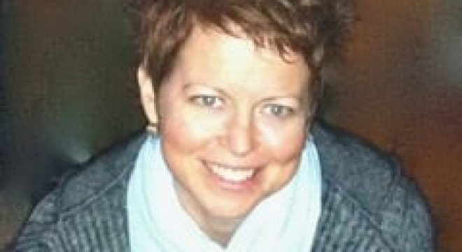 woman with short brown hair wearing a grey cardigan and light blue scarf smiles at the camera