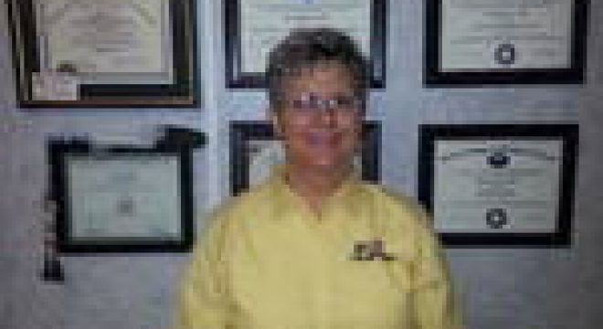 a woman in a yellow long-sleeve shirt smiles at the camera with six certificates hanging on the wall behind her